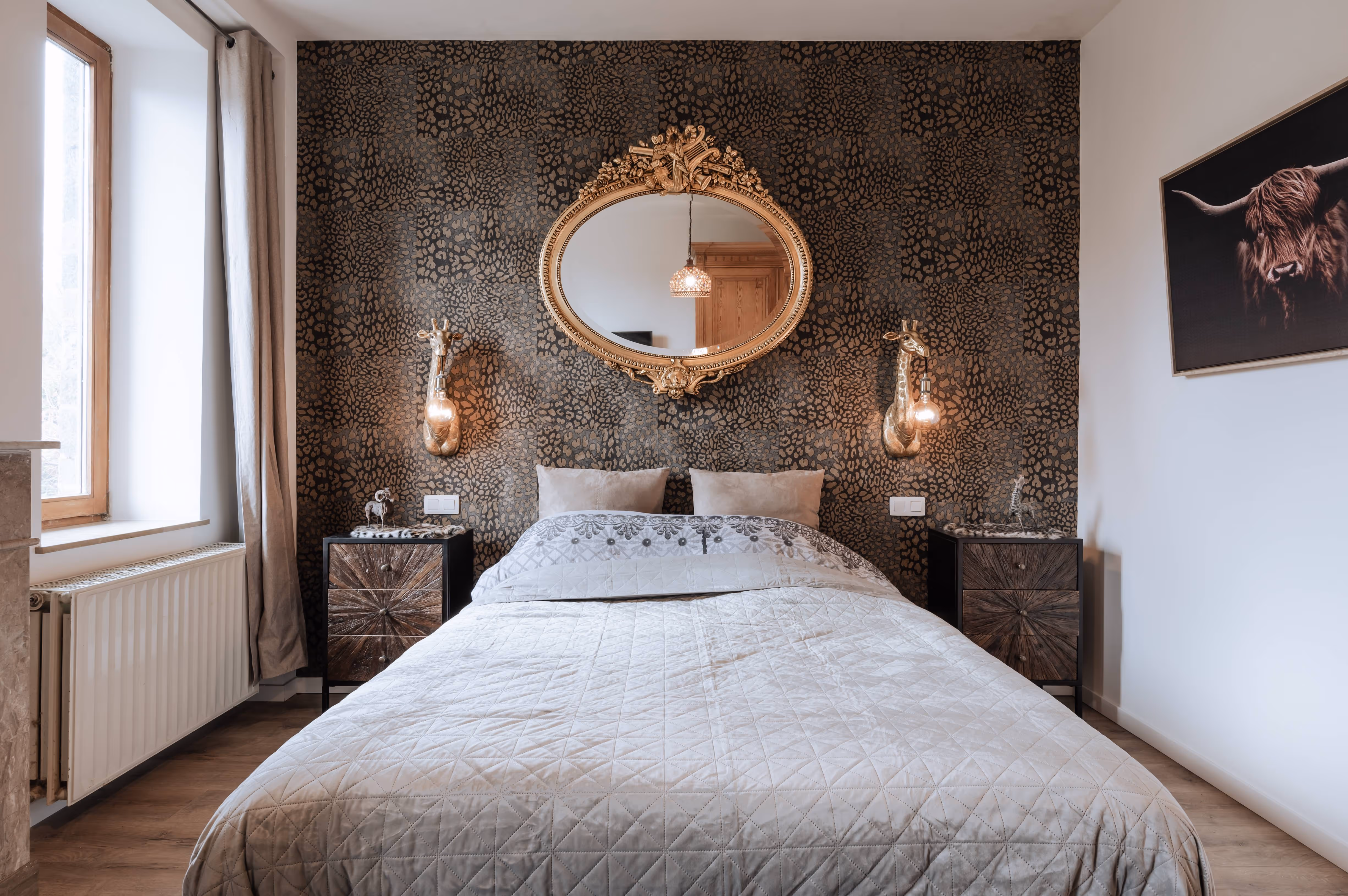 Bedroom with a patterned dark accent wall, gold oval mirror, giraffe-shaped wall lamps, a bed with beige bedding, two wooden nightstands, and a highland cow painting on the white wall.