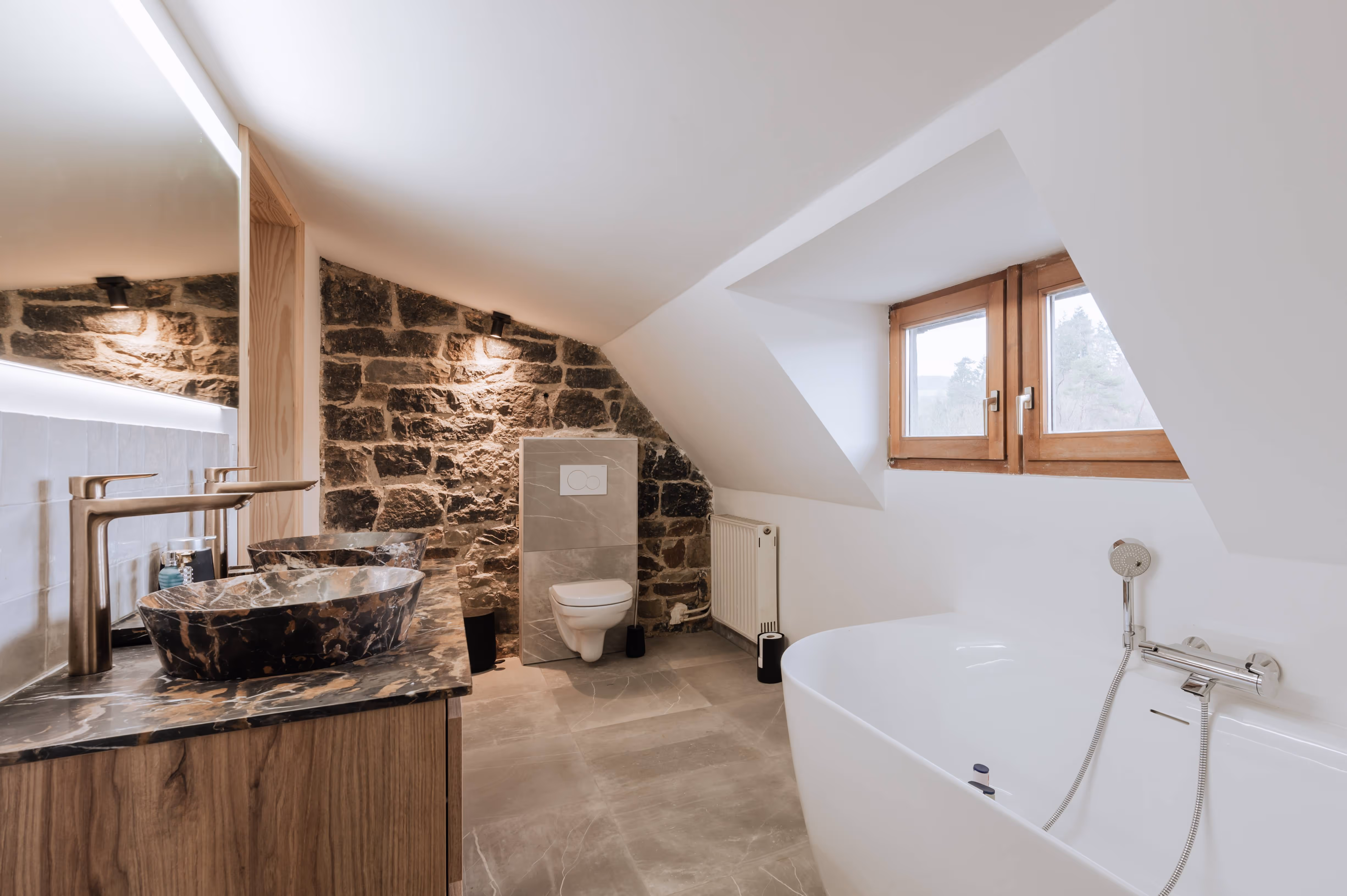 Modern bathroom with stone accent wall, marble countertop with vessel sinks, wall-mounted toilet, and freestanding bathtub under slanted ceiling.