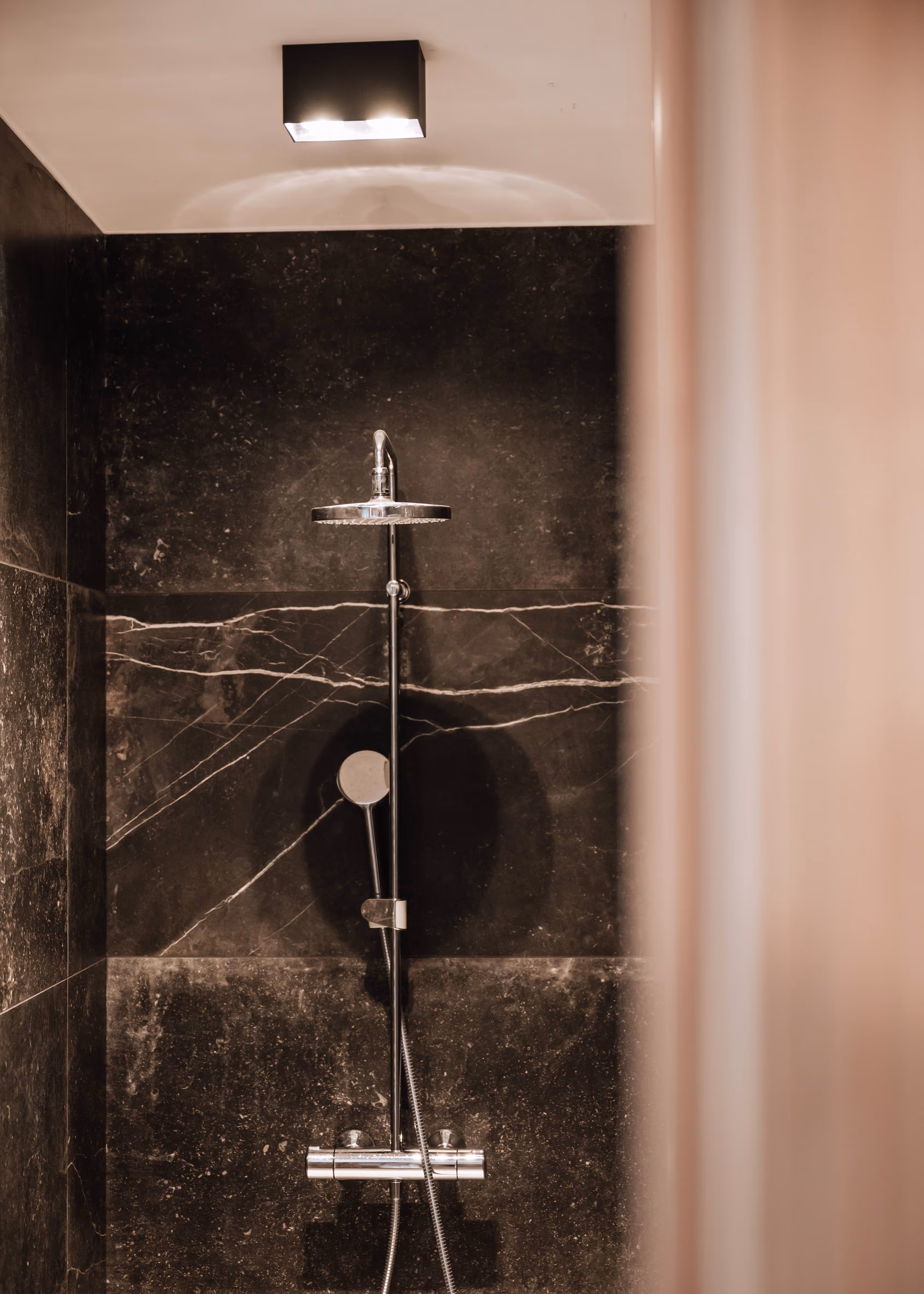 Modern shower with chrome fixtures mounted on dark marble walls under a ceiling light.