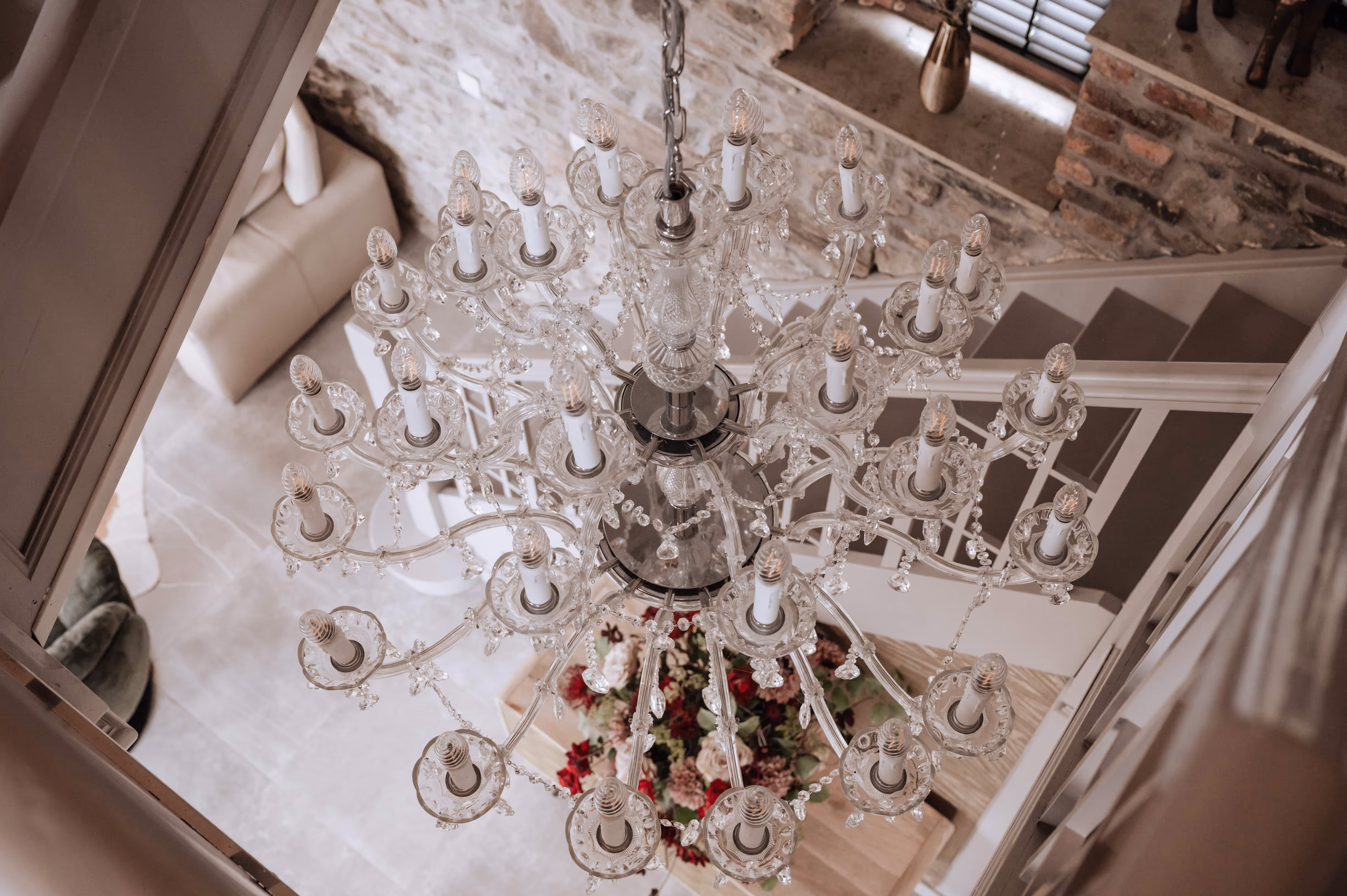 Elegant glass chandelier with multiple candle-shaped bulbs hanging above a staircase with a flower arrangement below.