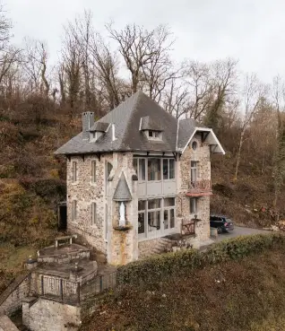 Stone house with steep roof and small balconies surrounded by leafless trees and bushes.