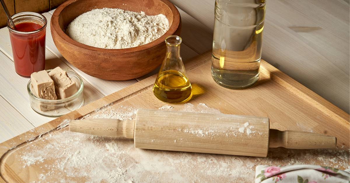 Ingrédients de base pour une pâte à pizza maison : farine blanche, huile d’olive extra vierge, levure fraîche, eau et sel. Préparation saine et artisanale sur plan de travail en bois.