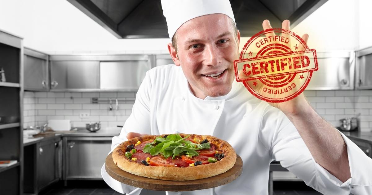 Pizzaiolo souriant tenant une pizza fraîche et un certificat de formation, ambiance cuisine professionnelle
