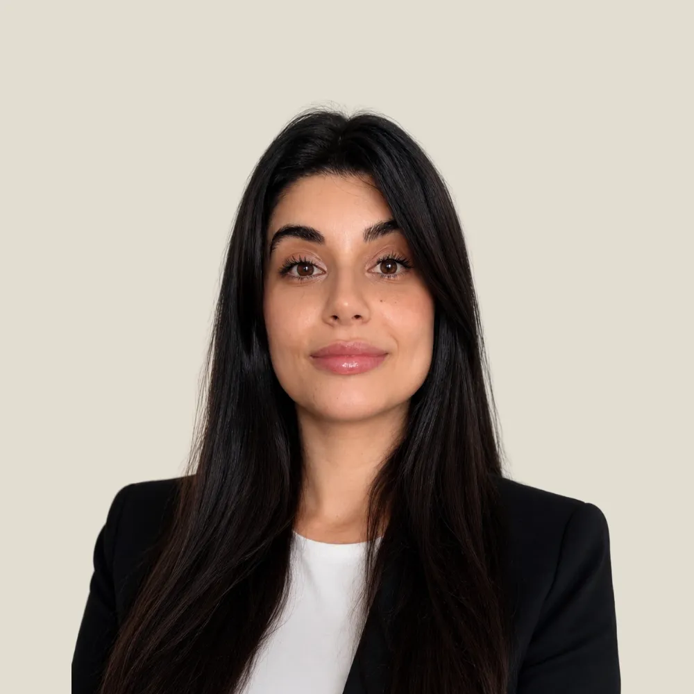 Portrait of a woman with long dark hair wearing a black blazer and white top against beige background.