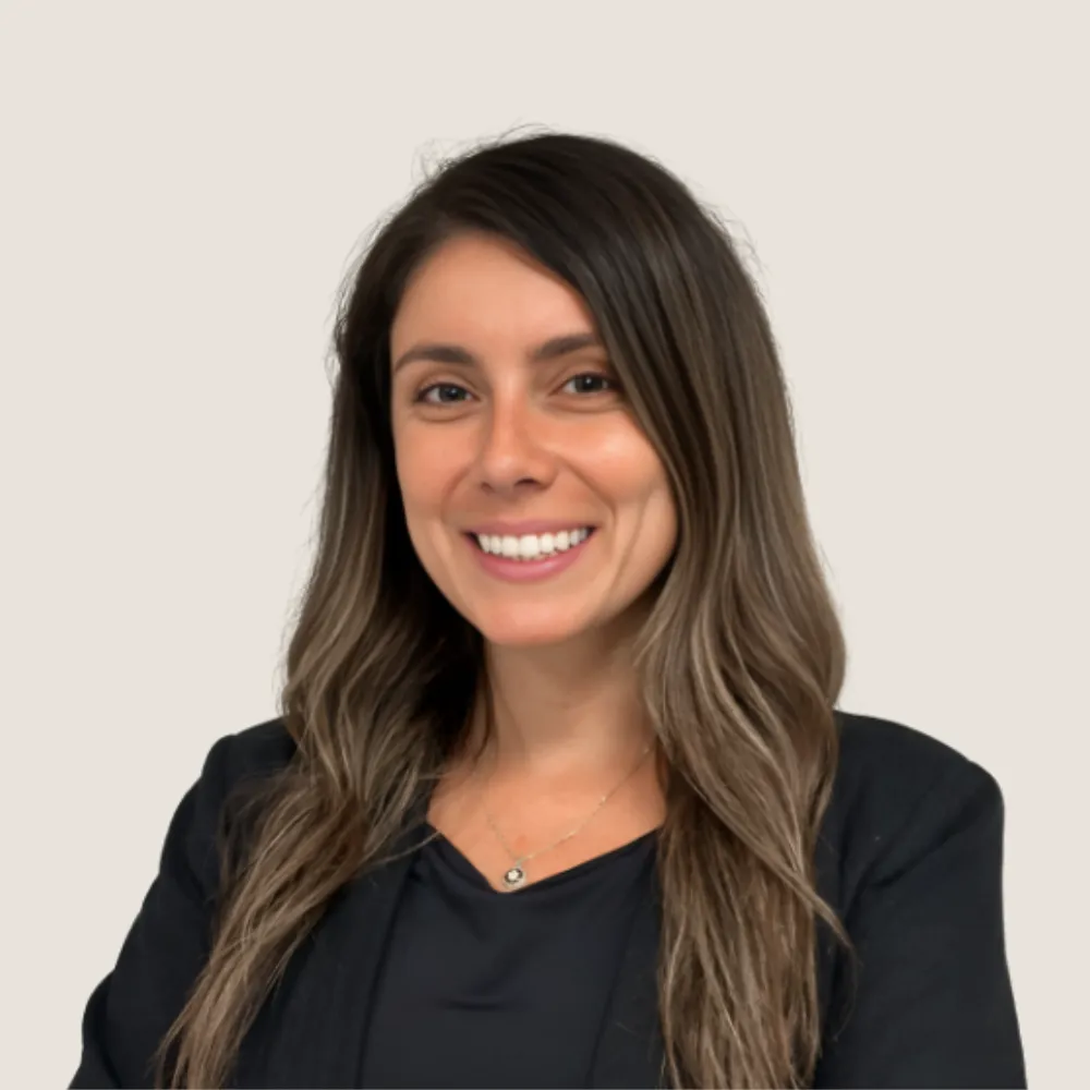 Smiling woman with long brown hair wearing a black blazer and necklace on a light background.