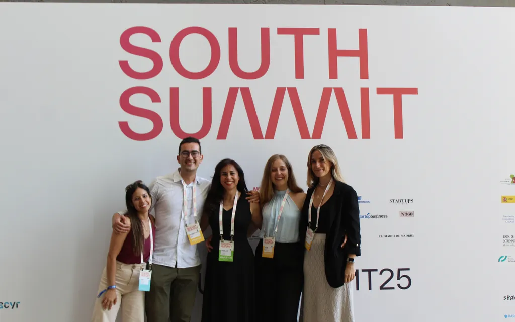 Five people smiling and posing in front of a white wall with large red South Summit text.