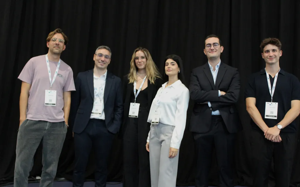 Group of six young professionals standing in front of a black curtain wearing conference badges.