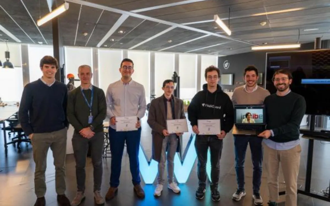 Seven men in an office, three holding certificates, one showing a laptop with a person on screen.
