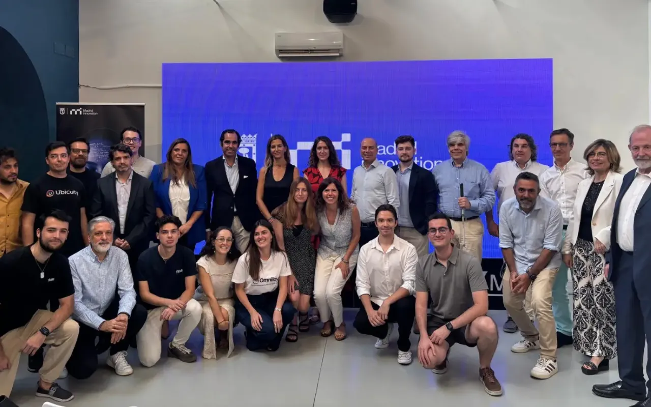 Group of 26 men and women posing in front of a blue screen inside an event space.