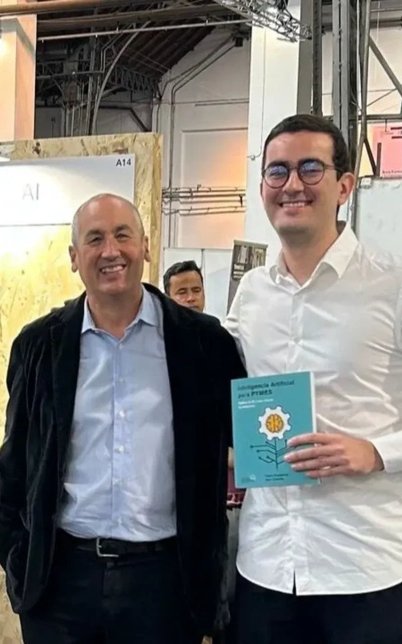 Two men smile indoors; the man on the right holds a book about artificial intelligence for SMEs.
