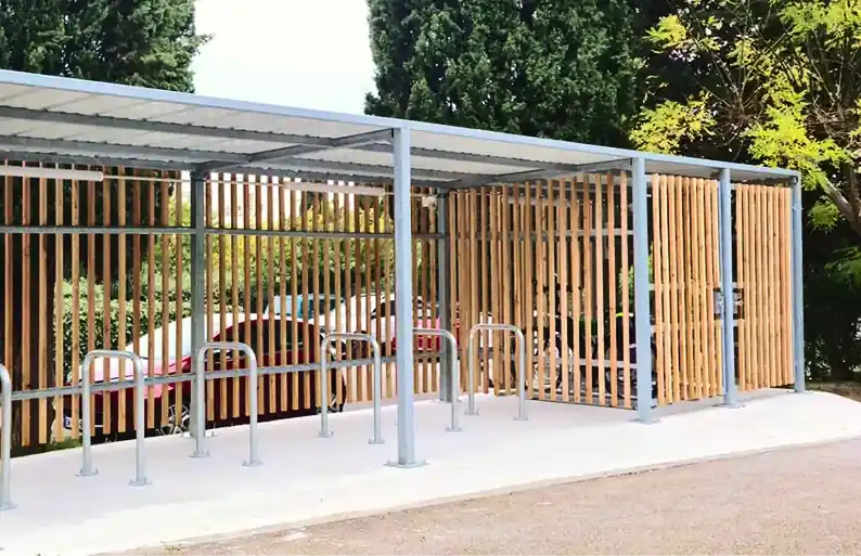 Abri en métal avec toit, équipé de supports métalliques pour vélos et de panneaux en lames de bois, entouré d’arbres verts.