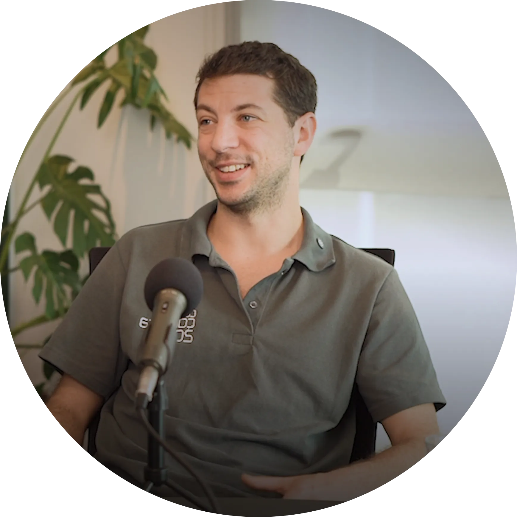 Hombre joven sentado frente a un micrófono, sonriendo con una planta al fondo.