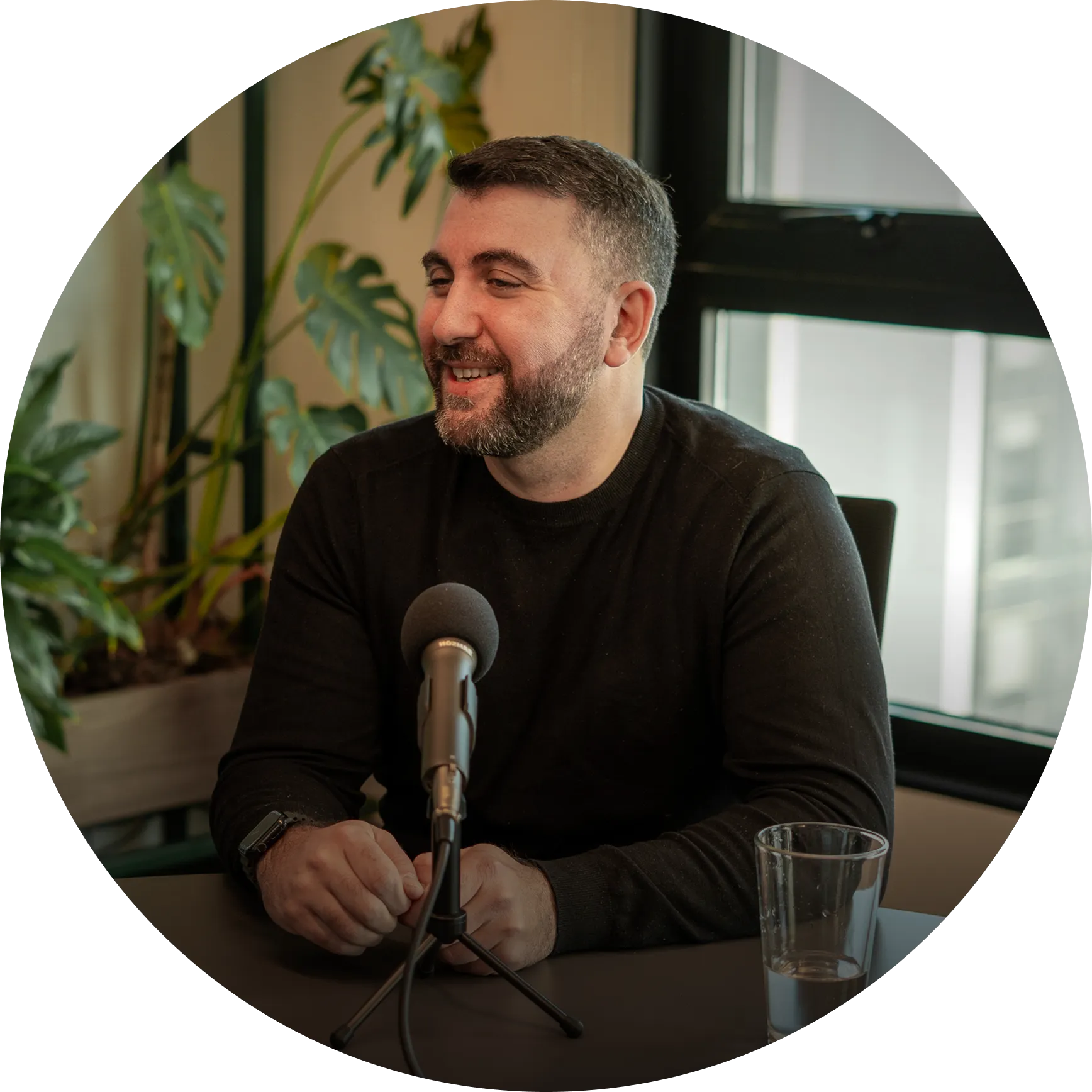 Mujer sonriente hablando frente a un micrófono en un entorno interior con plantas de fondo.