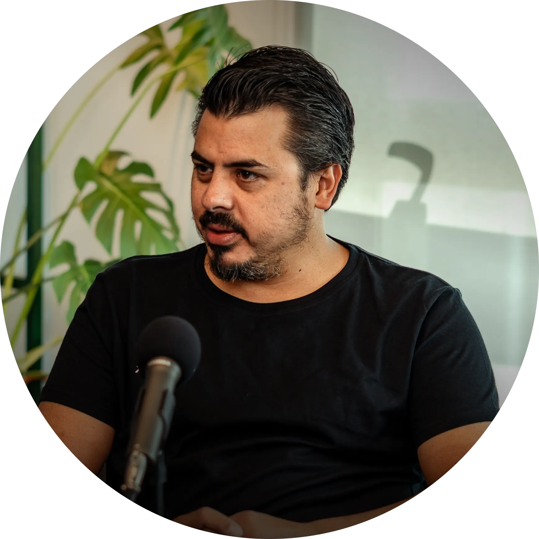 Hombre con barba y camiseta negra hablando frente a un micrófono con planta verde al fondo.