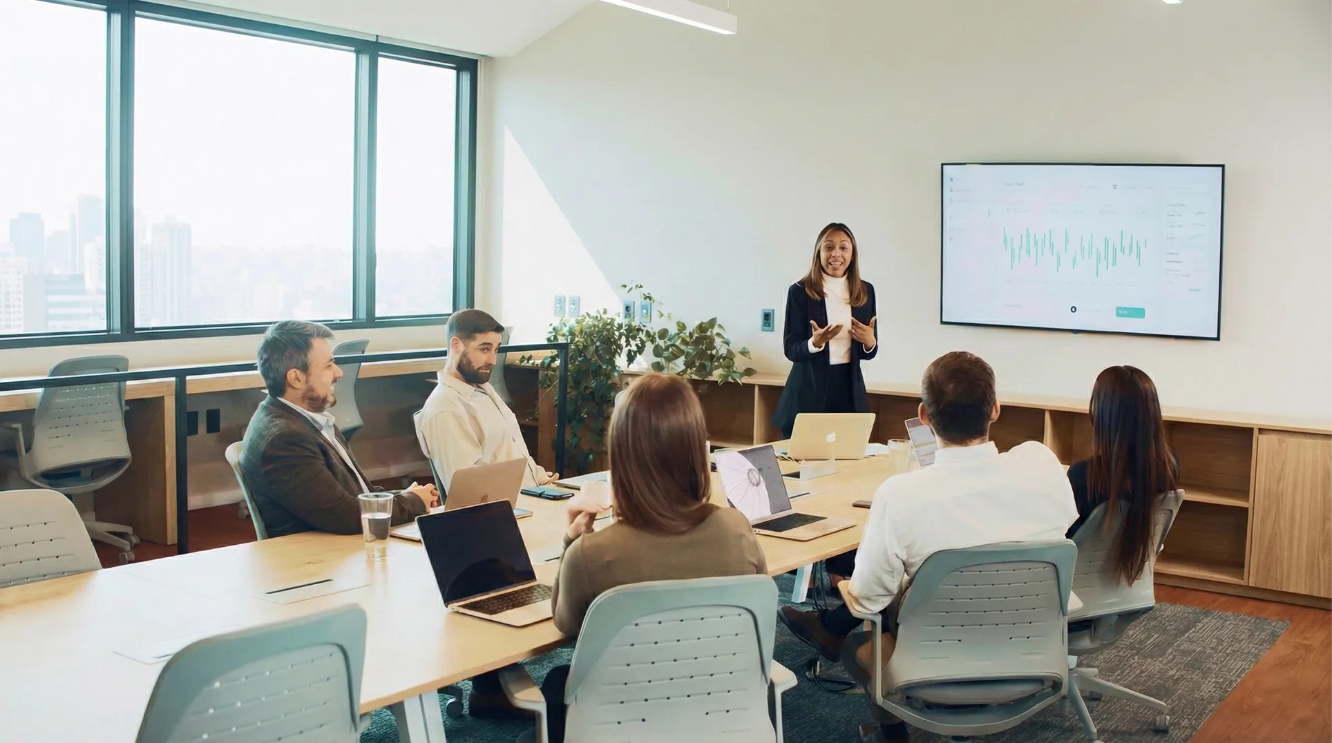 Mujer de negocios dando una presentación frente a una sala de reunión con cinco personas y una pantalla con gráficos.
