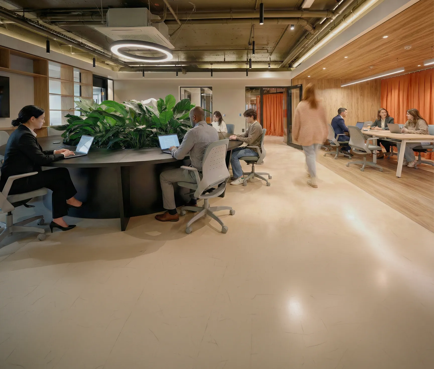 Personas trabajando con laptops en una oficina moderna de espacio compartido con plantas en una mesa central y paredes de madera con cortinas naranjas.