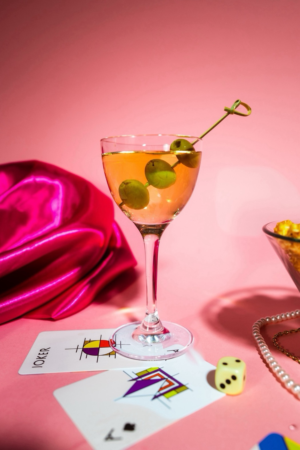 Glas mit Gin gefüllt, Spieß mit Olive, Spielkarten und eine Schüssel Popcorn, rosa Hintergrund