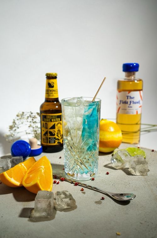 Eine Flasche First Flush Gin, ein Flasche Aqua Monaco, ein Longdrinkglas und Barutensilien, aufgeschnittene Orange und Eiswürfel