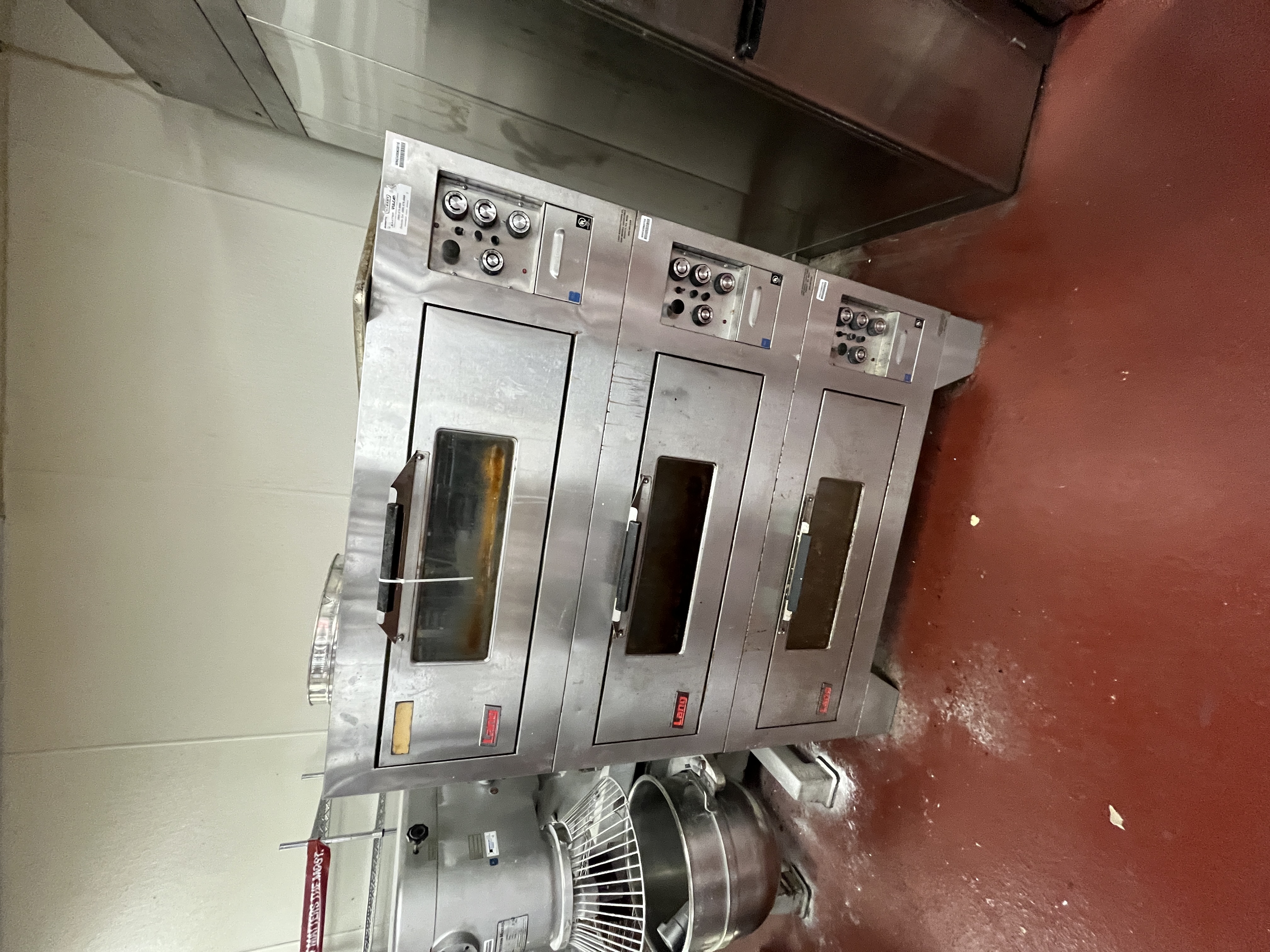 Stainless steel triple-stack Lang commercial bakery oven with viewing windows and control panels, located in a professional grocery store bakery setting. High-quality commercial cooking equipment available from Mid-America Store Fixtures.