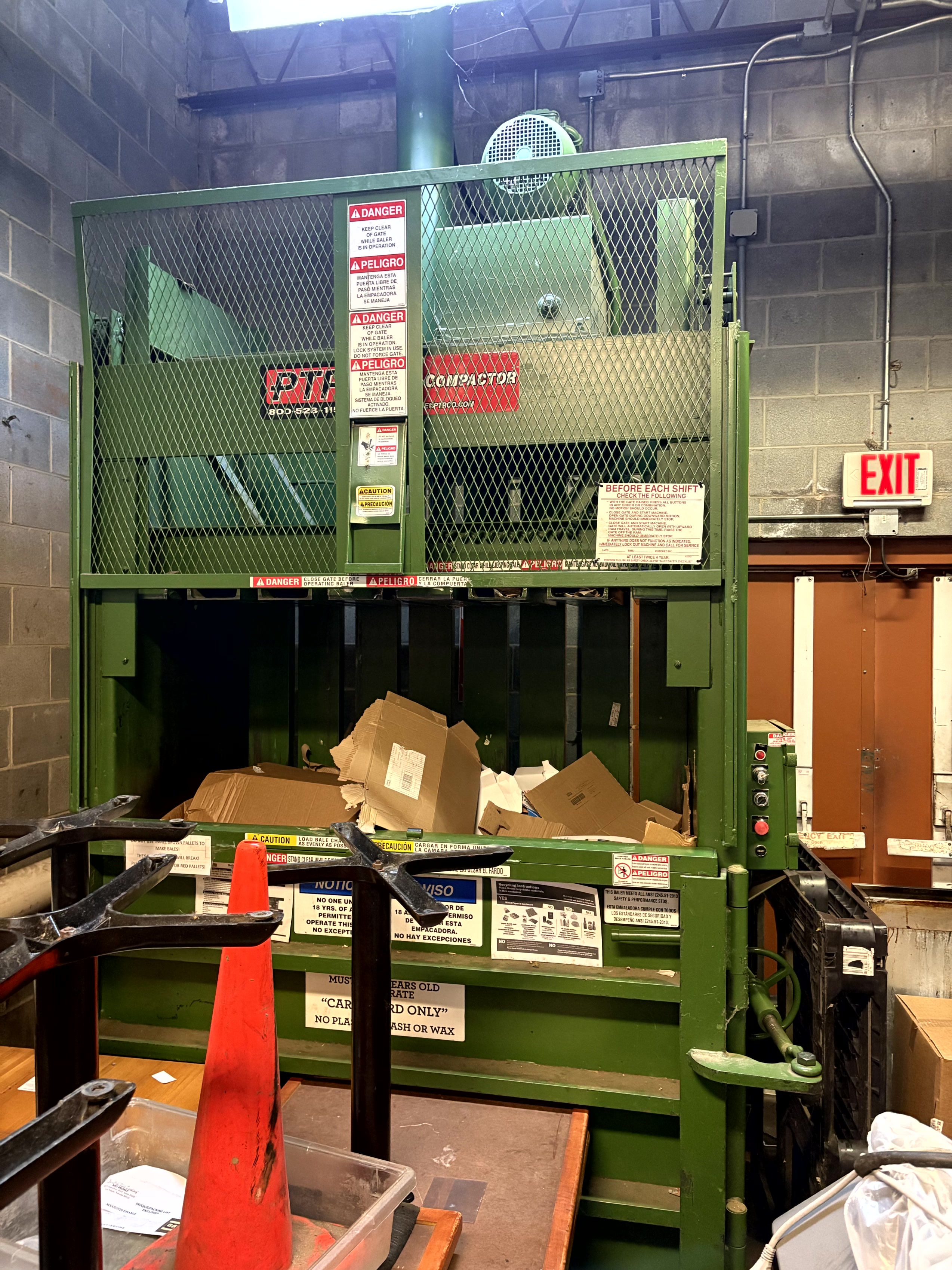 Green PTR cardboard baler and compactor machine in a commercial backroom area, used for compressing cardboard waste and recycling materials. Industrial baler equipment available from Mid-America Store Fixtures.