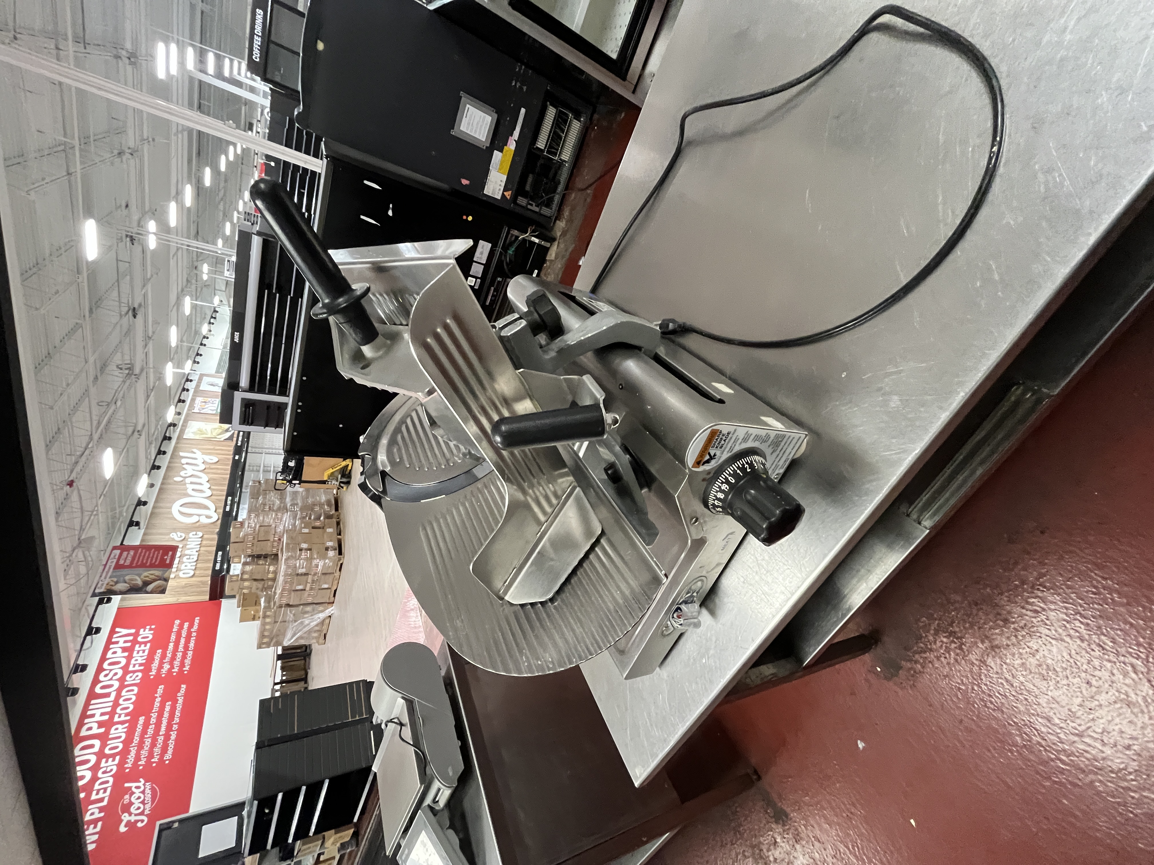 Commercial deli meat slicer on stainless steel prep table inside a grocery store. Professional slicer available from Mid-America Store Fixtures.