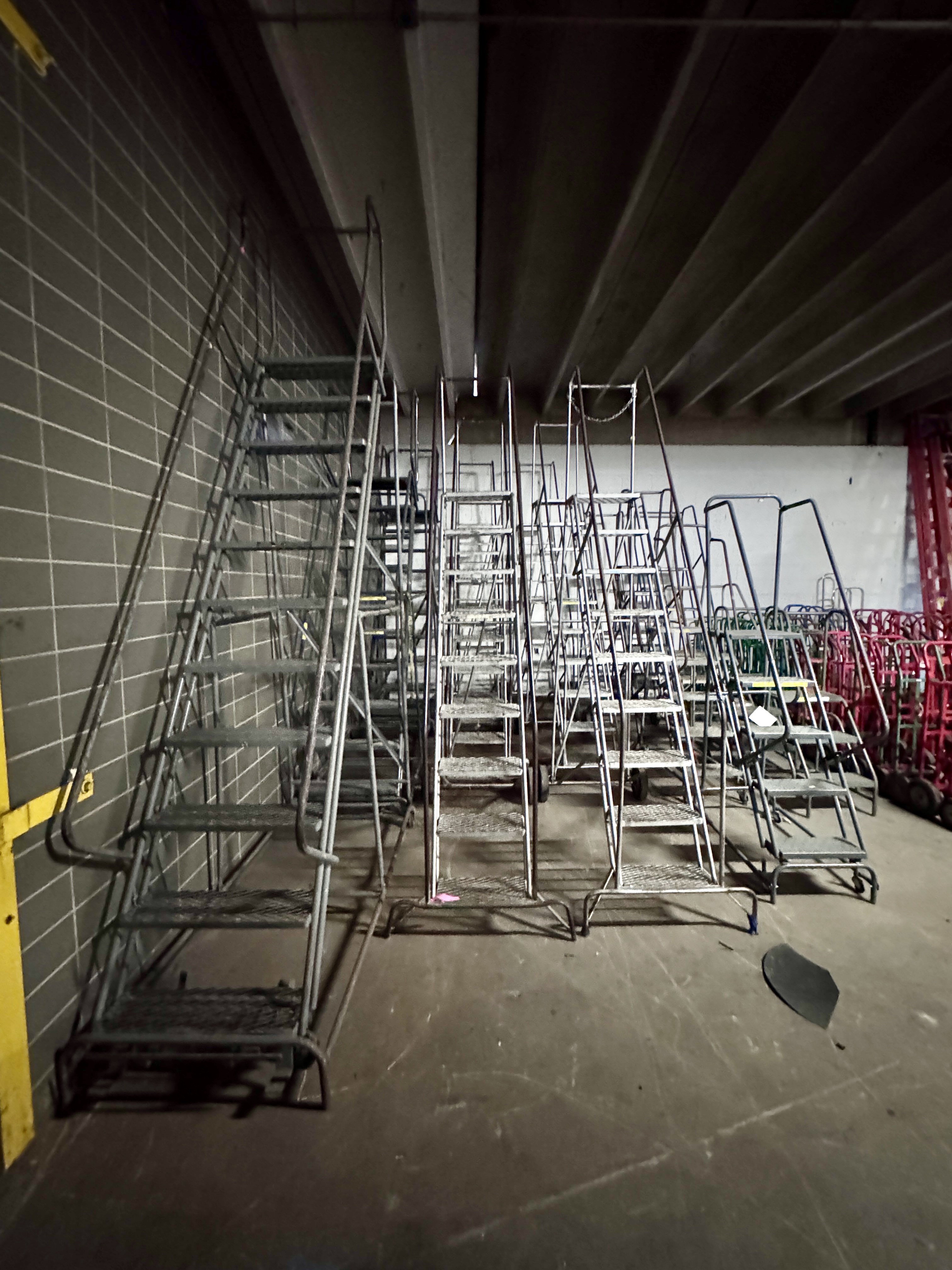 Row of industrial rolling safety ladders with handrails and slip-resistant steps used in warehouse and stockroom environments. Heavy-duty rolling ladders available from Mid-America Store Fixtures.