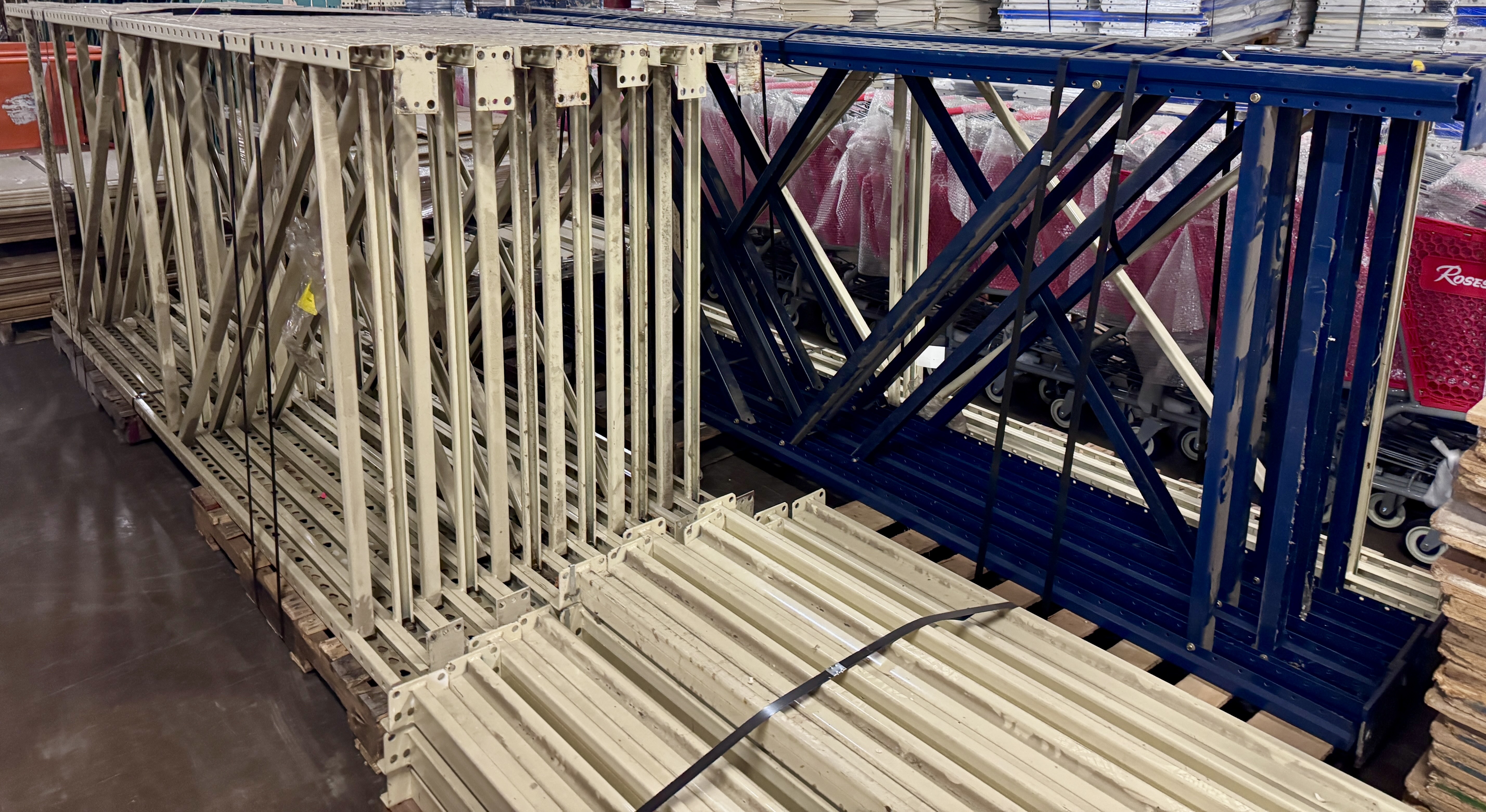 Bundles of tan and blue teardrop pallet racking uprights stacked on pallets inside a warehouse. Heavy-duty industrial rack uprights available from Mid-America Store Fixtures.