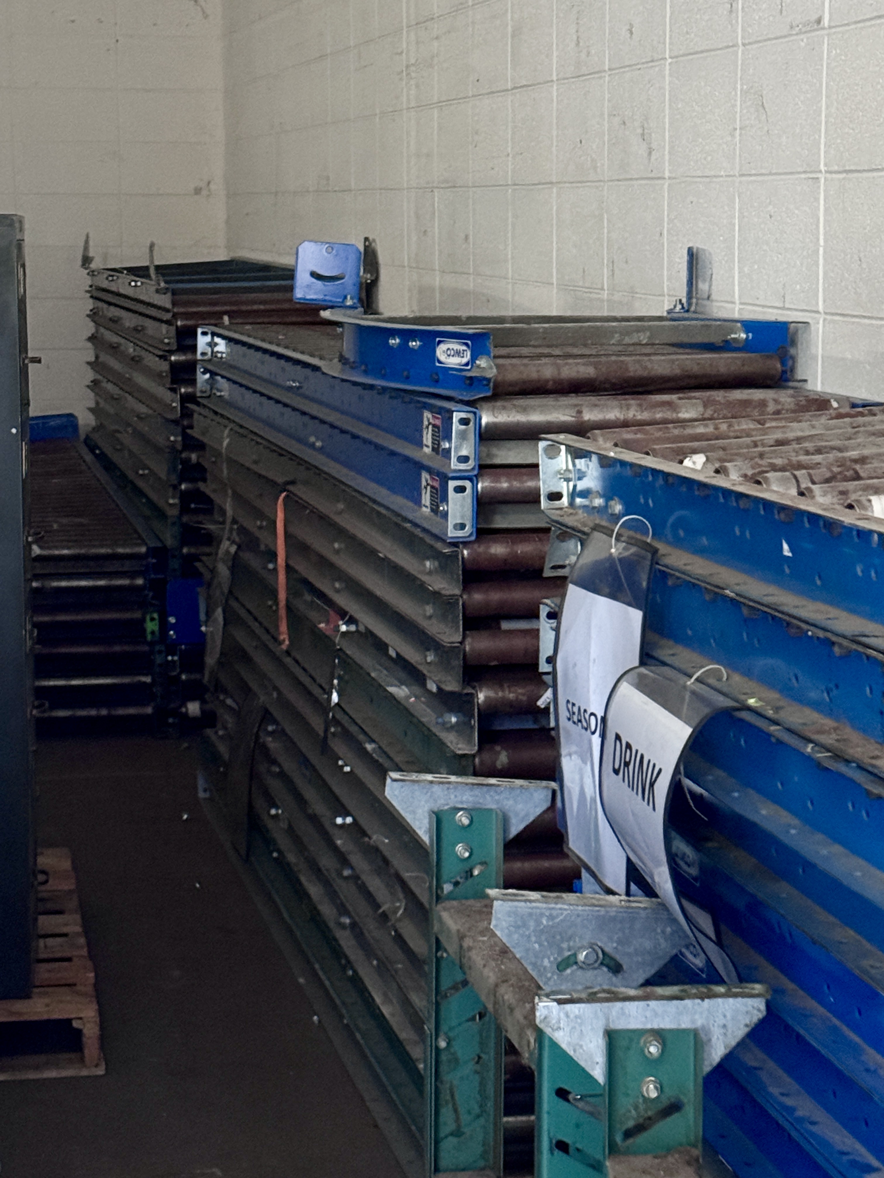 Stacks of blue and green industrial roller conveyor sections stored in a warehouse, ideal for stockrooms, distribution centers, and material handling lines. Roller conveyor equipment available from Mid-America Store Fixtures.