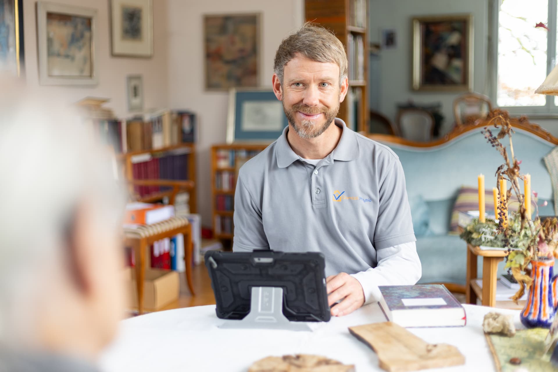 Physiotherapeut sitzt mit einem Tablet am Wohnzimmertisch und bespricht die Behandlung während eines Hausbesuchs.