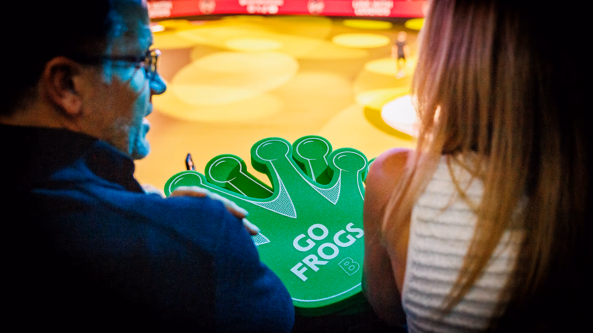 Two people holding green foam clappers shaped like a frog foot with text 'GO FROGS' at a sports or event venue.