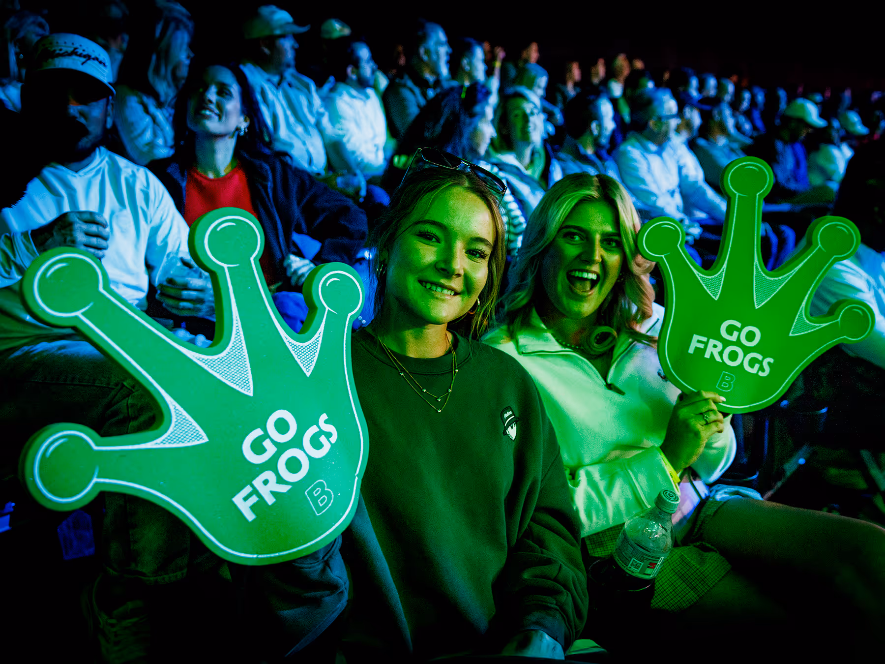 Two women at an event holding large green foam frog fingers with text 'GO FROGS' and smiling among a crowd.