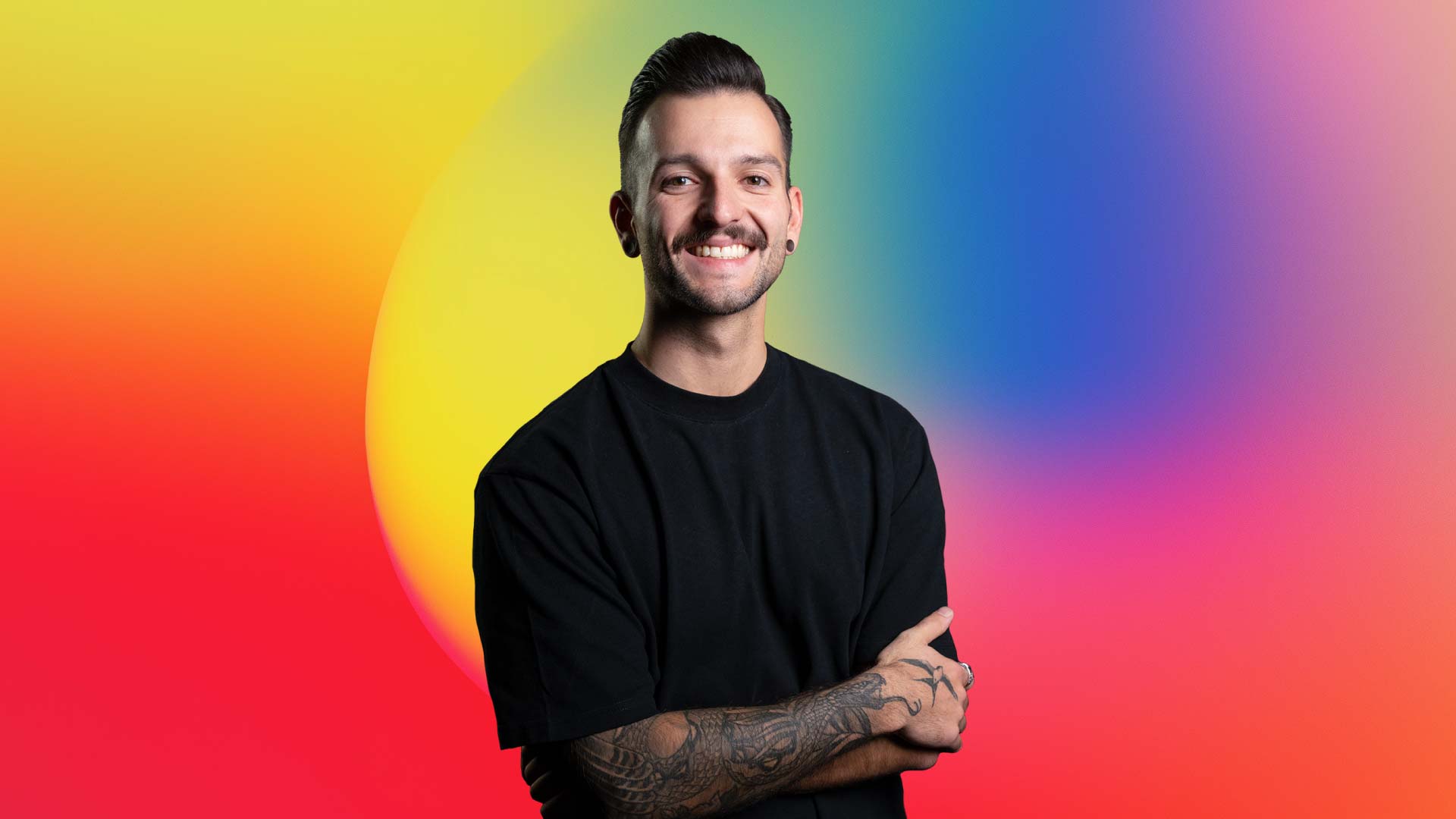 Smiling man with tattooed forearm and black shirt standing against a colorful gradient background.
