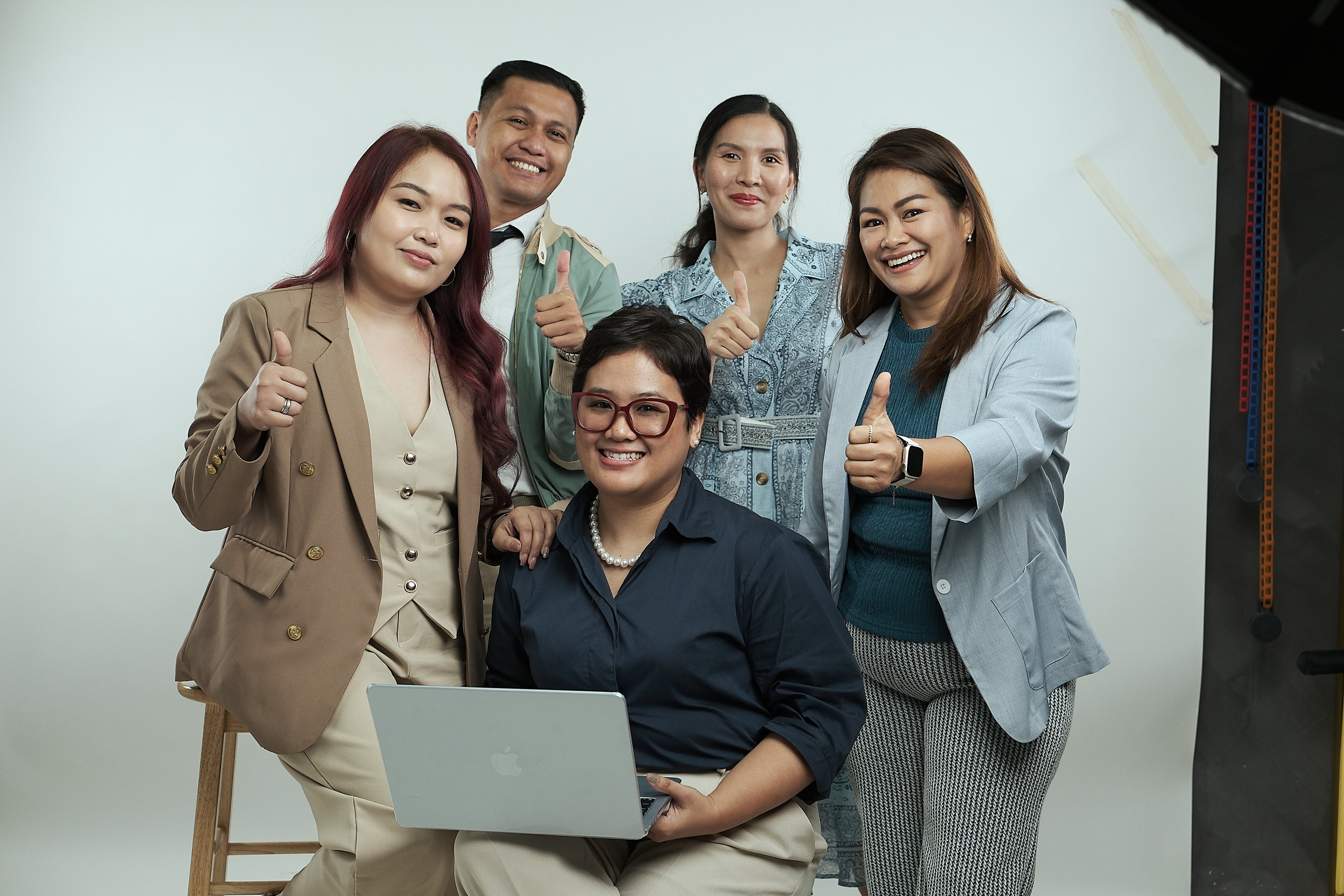 Five diverse professionals smiling and giving thumbs-up, one seated with a laptop. | Simple for clients. 
Meaningful for the people doing the work. | RemoteGenies