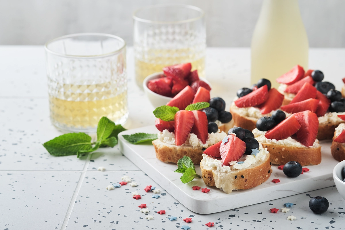Fresh berry bruschetta with strawberries, blueberries, whipped cream, and mint served alongside juice glasses and patriotic red, white, and blue sprinkles in a bright luxury Fourth of July brunch setting in Scottsdale