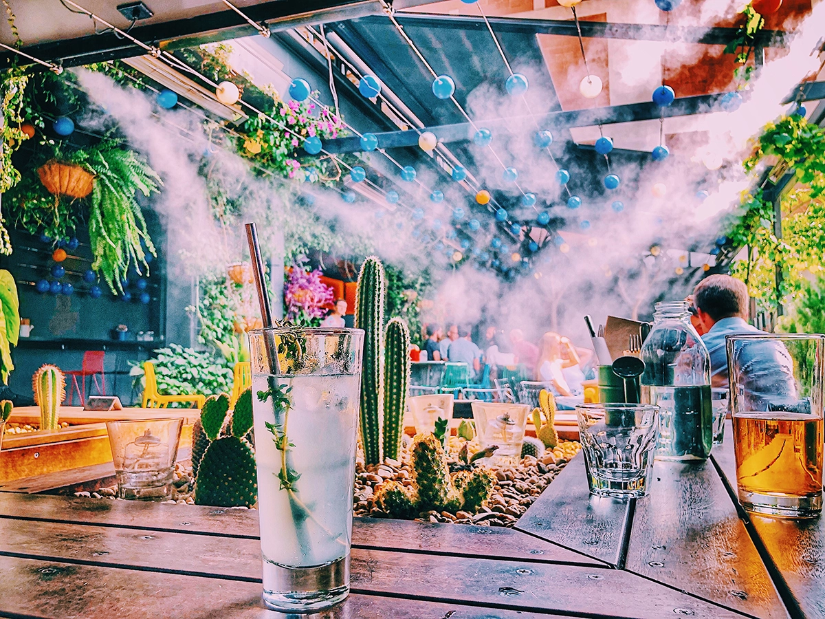 Guests relaxing at a Scottsdale luxury event comfort station with infused water, misting, rustic wooden tables, and lush garden-inspired décor beneath a glass ceiling