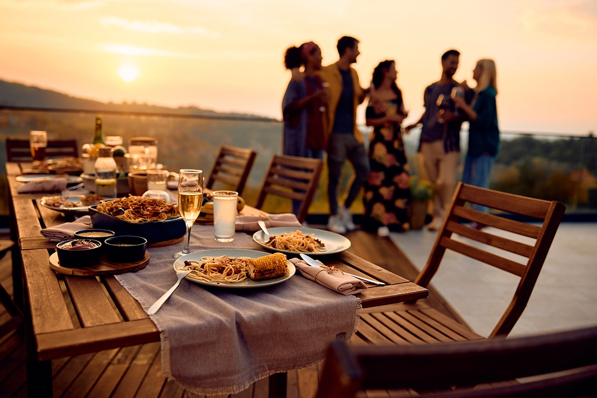 A wooden table set for an outdoor dinner on a terrace at dusk, bathed in the warm glow of the setting sun. Plates of pasta and corn, along with various dips and drinks, are arranged across the table, while several casually dressed adults stand in the backg