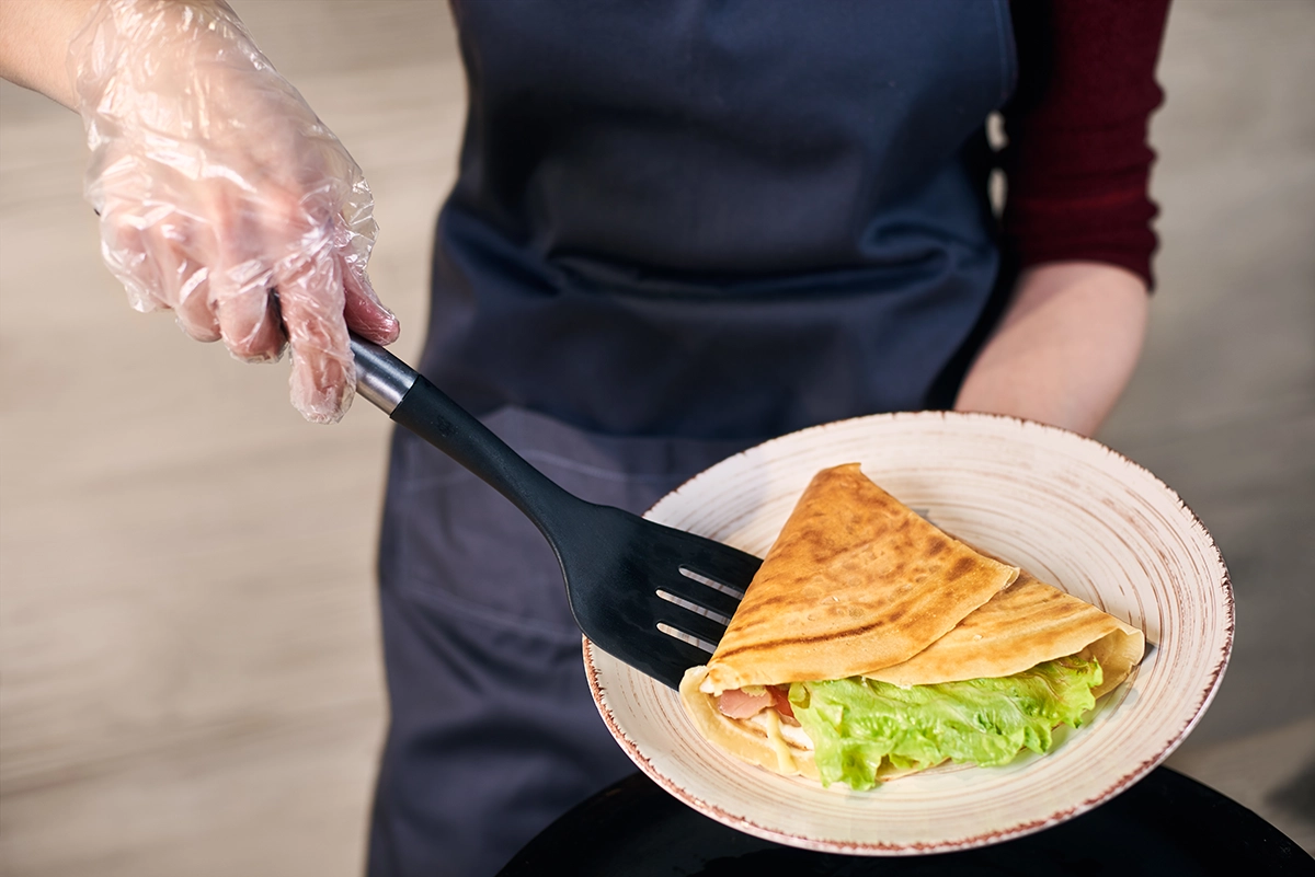 A chef in an apron and gloves uses a spatula to serve a freshly made crepe folded around leafy greens, white sauce, and sliced meat at a live brunch station.
