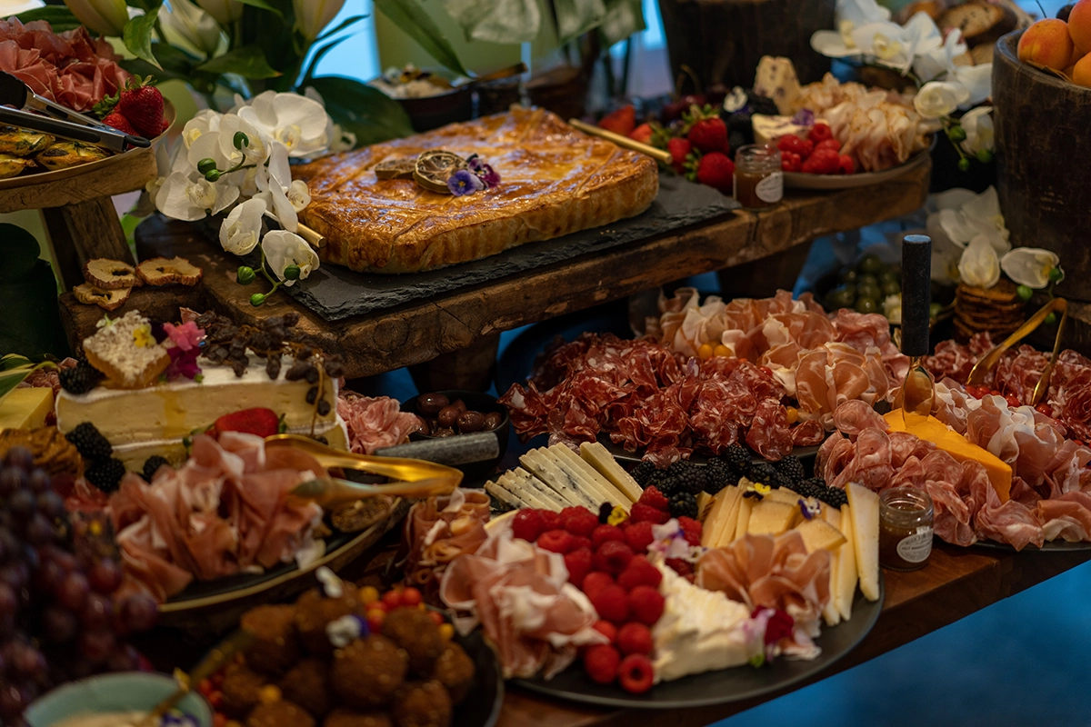 An abundant brunch grazing table featuring a charcuterie board of cheeses, cured meats, and fresh fruit, with a golden-brown pastry and flowers in a wooden bucket, arranged on a tiered wooden display for a special event.