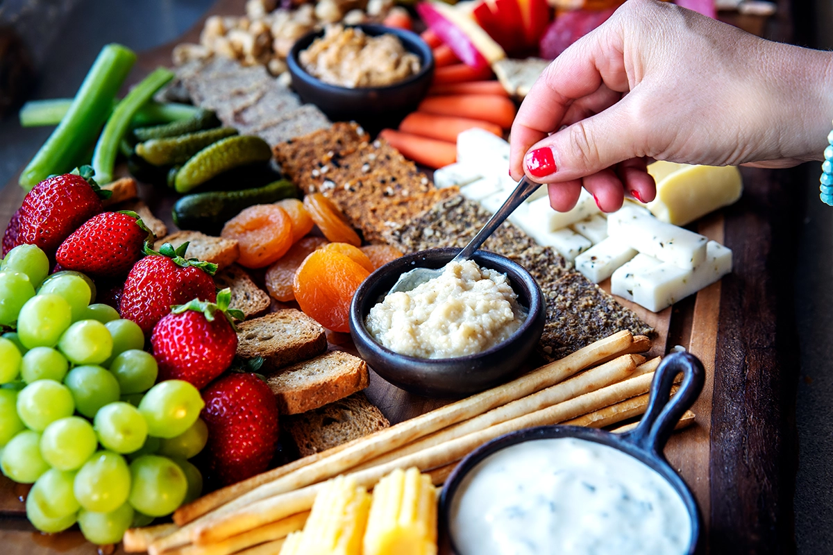 Shared grazing board with cheeses, cured meats, fresh fruit, breads, crackers, and dipping sauces on an office table, with guests reaching in during a team lunch.