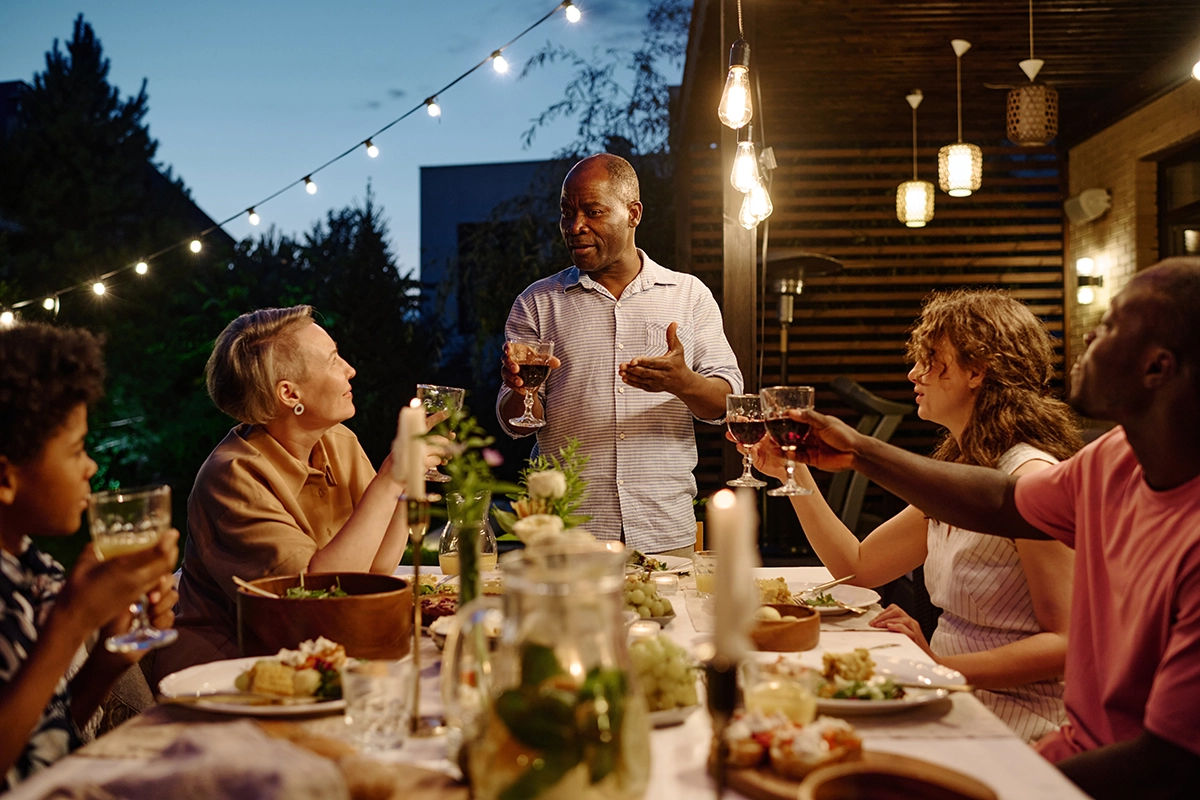 Six guests, including a child, sharing a meal at an outdoor table lit by hanging lights at night.