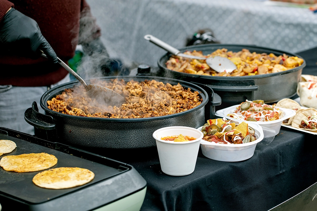 A gloved food service worker stirring a steaming pot beside corn tortillas, bowls of nachos, and cups of soup at a taco station.
