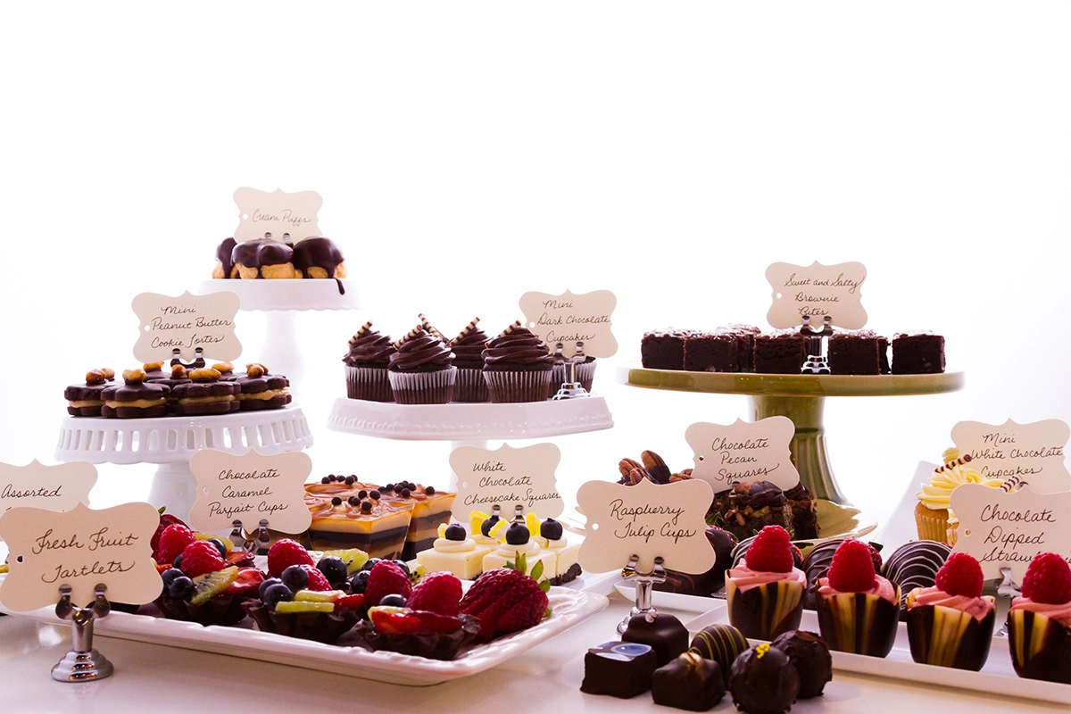 A dessert table of individually portioned brownies, chocolate cheesecake squares, fruit tarts, and cupcakes arranged on stands with labels.