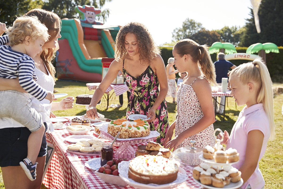 An adult serving slices of cake to children at an outdoor party table of desserts, with another adult holding a small child nearby.