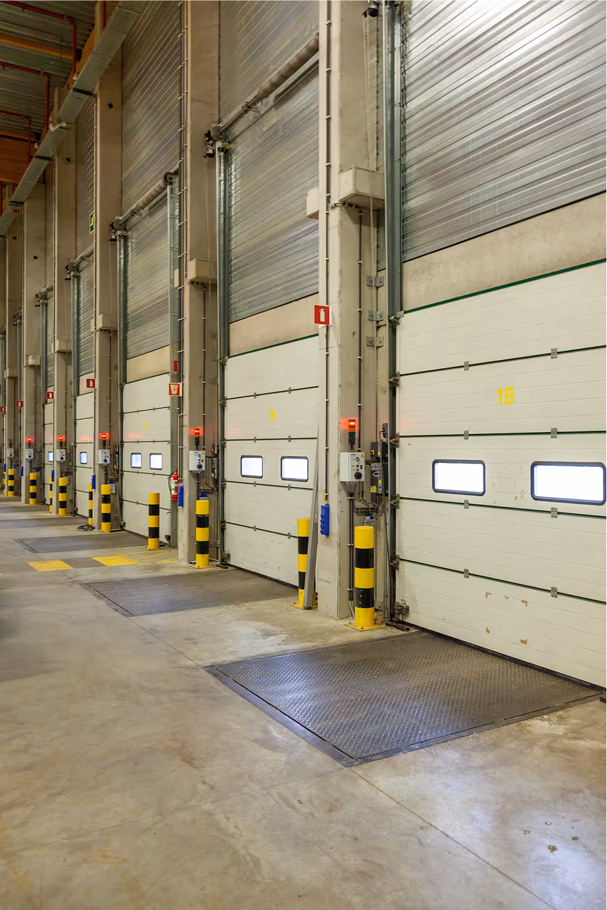 Interior of a warehouse loading area with multiple closed white roll-up doors numbered and protected by yellow-black striped bollards.