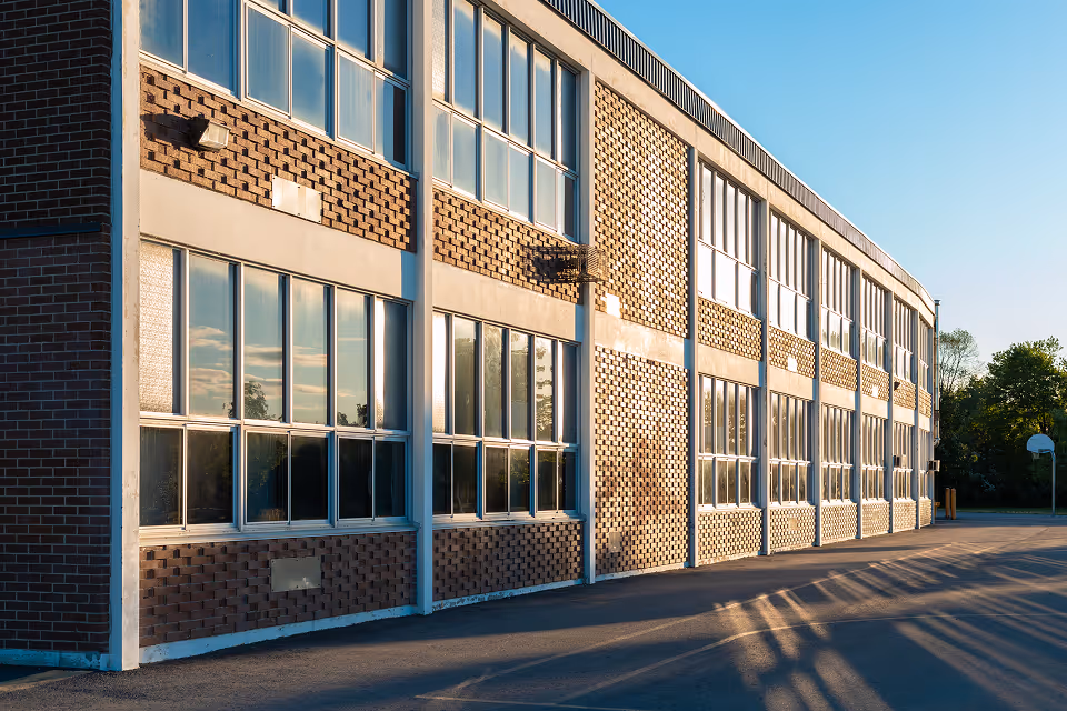 Sunlit brick school building with large windows and a basketball hoop on the right side.