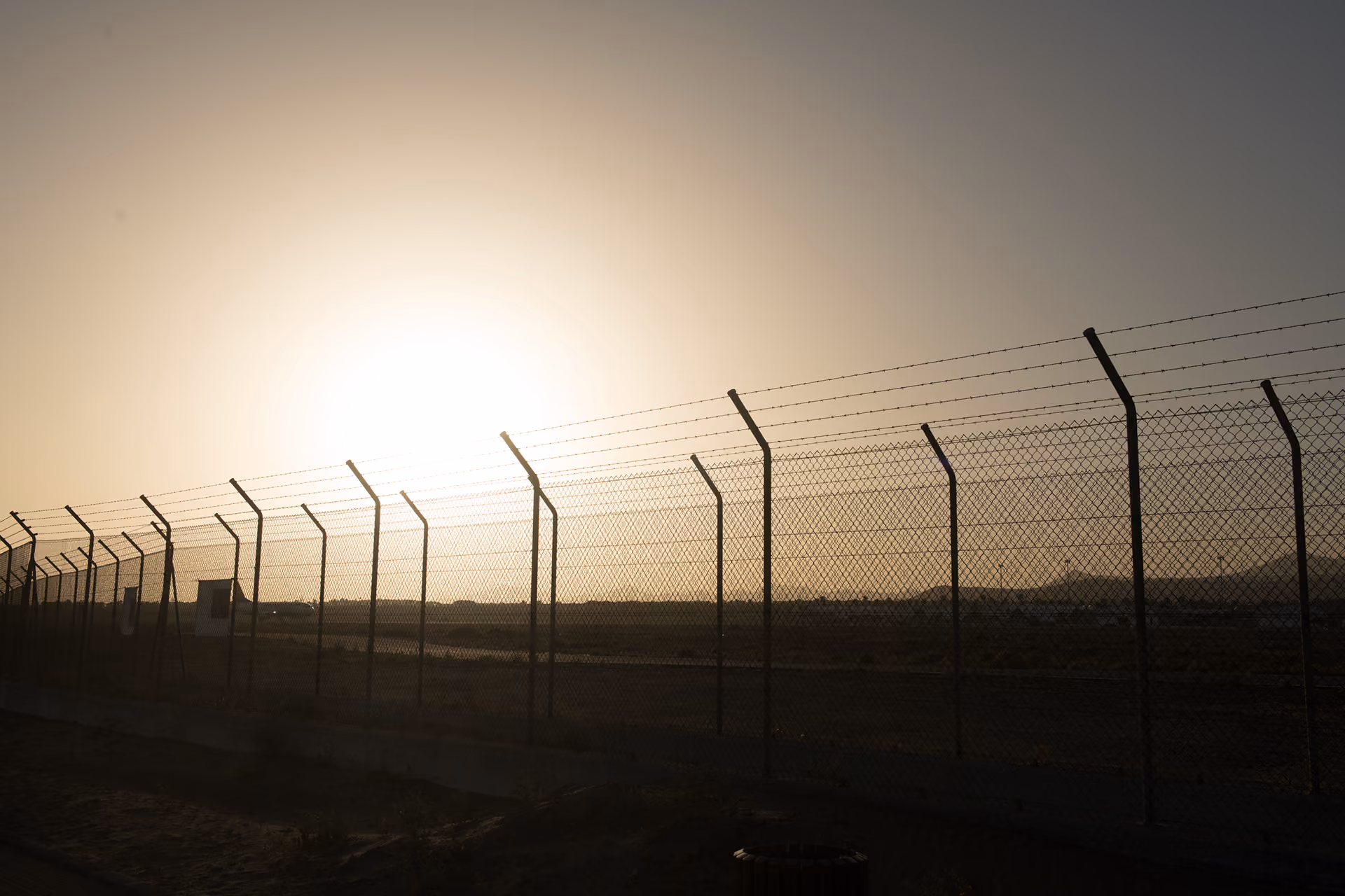 Long chain-link fence topped with barbed wire at sunset over a barren landscape.