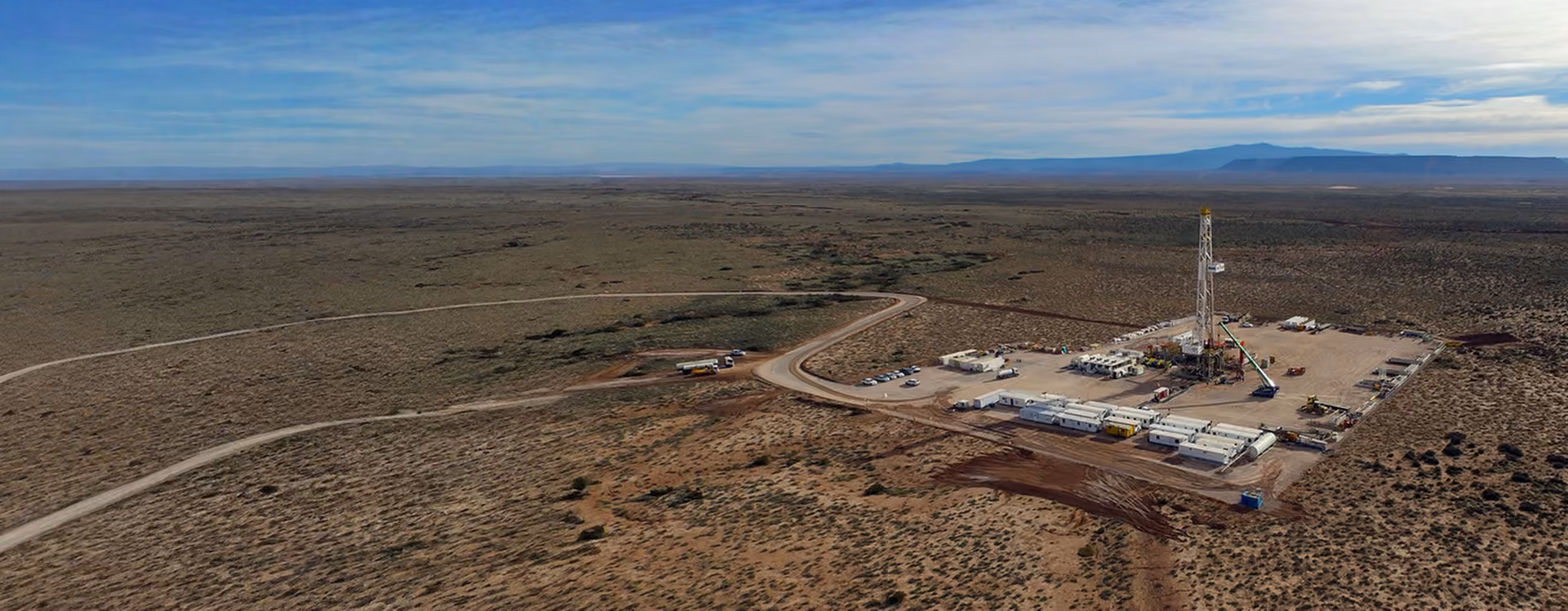 Remote drilling site with equipment and vehicles in a vast, dry, flat landscape under a blue sky.