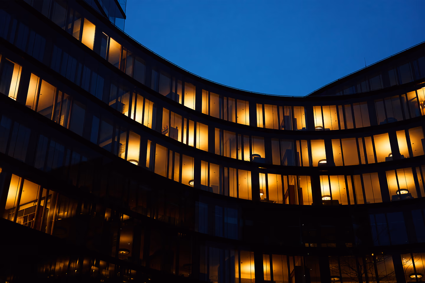 Curved office building at dusk with warmly lit interior rooms visible through glass windows.