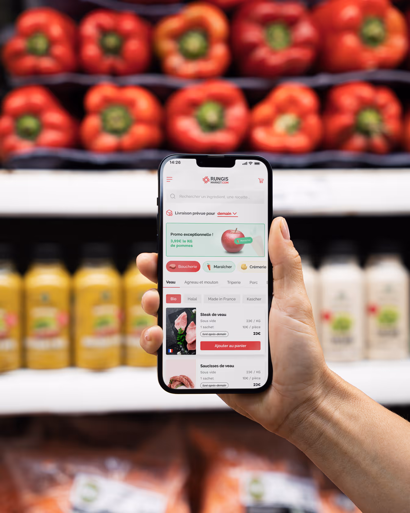Main tenant un smartphone ouvert sur l’application Rungis Market devant un rayon de poivrons rouges en supermarché.