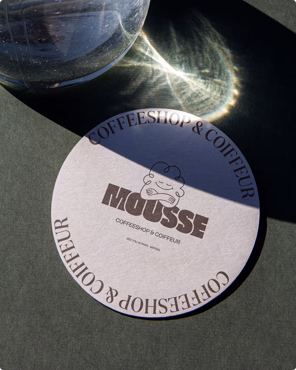 Dessous de verre rond avec le logo Mousse coffeeshop & coiffeur imprimé en brun posé sur une table verte.